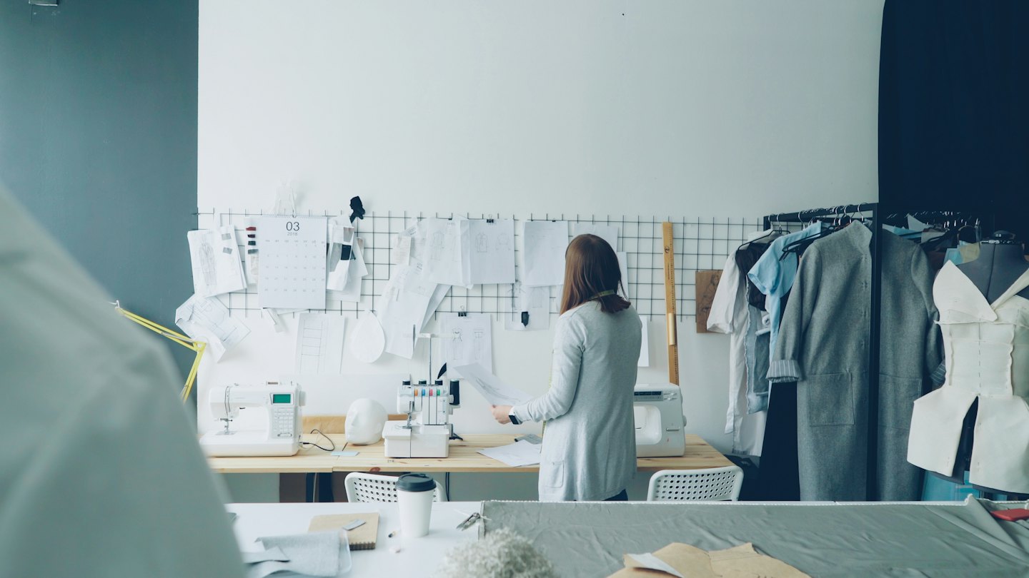 Fashion designer reviews patterns at a wooden workbench in her studio.